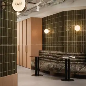 Góc ngồi quán cà phê sử dụng gạch subway màu xanh olive men bóng kích thước khoảng 75x300 mm, phối nội thất gỗ sáng và đèn tường phong cách tối giản.
